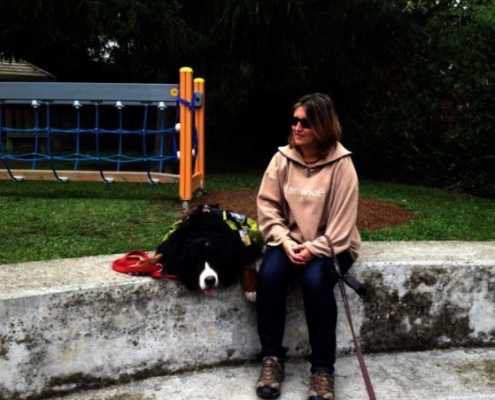 Une dame assise sur un muret aux côtés de son chien d’assistance Babou, couché près d’un terrain de jeux extérieurs.