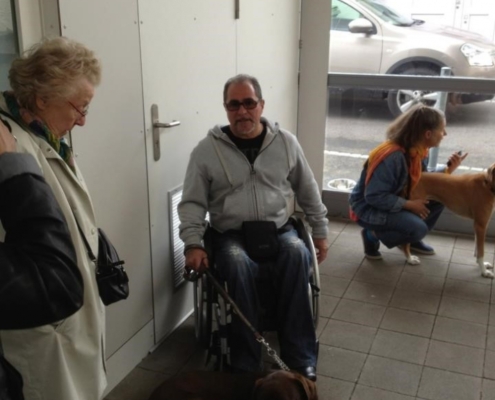 Une dame en manteau clair avec un homme en fauteuil et son chien d’assistance, jeune dame à droite avec un second chien.