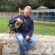 Un homme assis caressant un petit chien noir en manteau, dans une aire de jeux.