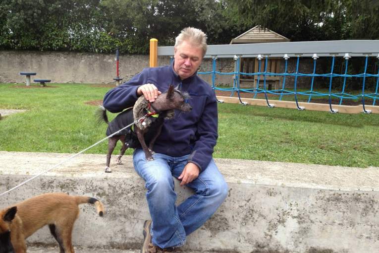 Un homme assis caressant un petit chien noir en manteau, dans une aire de jeux.