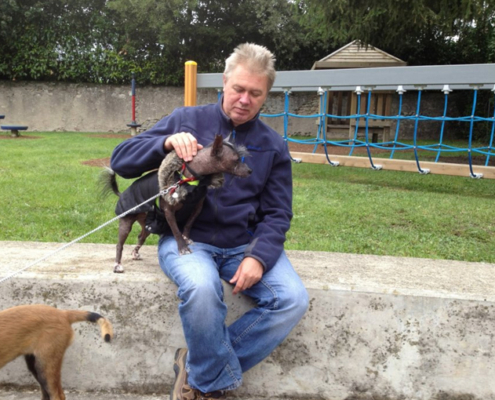 Un homme assis caressant un petit chien noir en manteau, dans une aire de jeux.