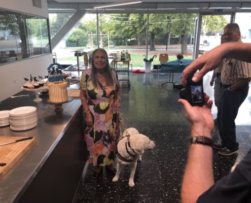 Dame souriante posant près d’un gâteau d’anniversaire avec son chien guide allongé, tandis qu’une personne prend une photo.