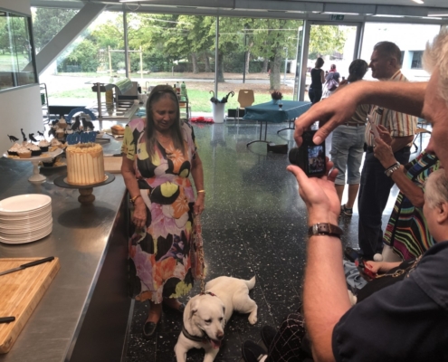 Dame souriante posant près d’un gâteau d’anniversaire avec son chien guide allongé, tandis qu’une personne prend une photo.