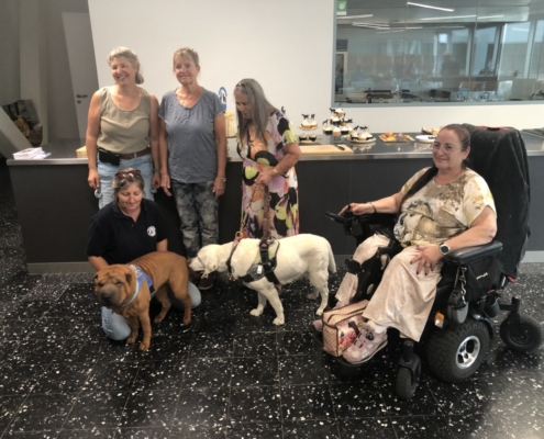Photo de groupe avec cinq dames et deux chiens d’assistance devant un comptoir garni de gâteaux et cupcakes.