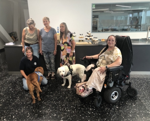 Photo de groupe avec cinq dames et deux chiens d’assistance devant un comptoir garni de gâteaux et cupcakes.
