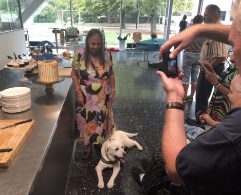 Dame souriante posant près d’un gâteau d’anniversaire avec son chien guide allongé, tandis qu’une personne prend une photo.
