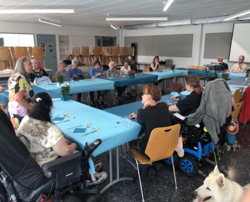 Une dame debout s’adressant aux invités d’une fête conviviale autour de grandes tables bleues, en présence de chiens d’assistance.