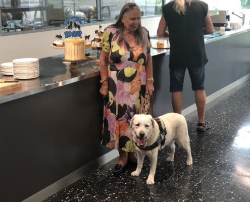 Une dame debout avec un chien guide devant un gâteau d’anniversaire décoré « 20 ans », sur un buffet dans une salle lumineuse.