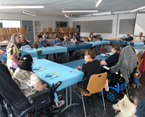 Une dame debout s’adressant aux invités d’une fête conviviale autour de grandes tables bleues, en présence de chiens d’assistance.