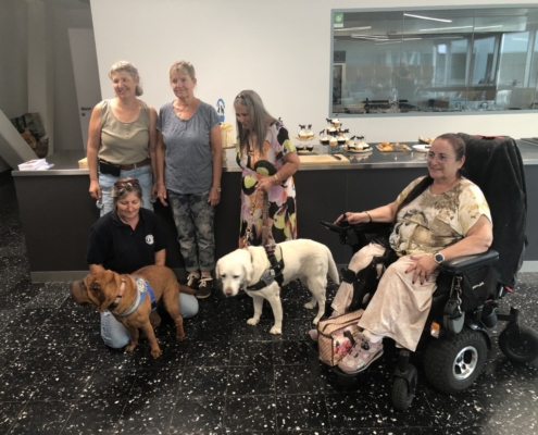 Photo de groupe avec cinq dames et deux chiens d’assistance devant un comptoir garni de gâteaux et cupcakes.