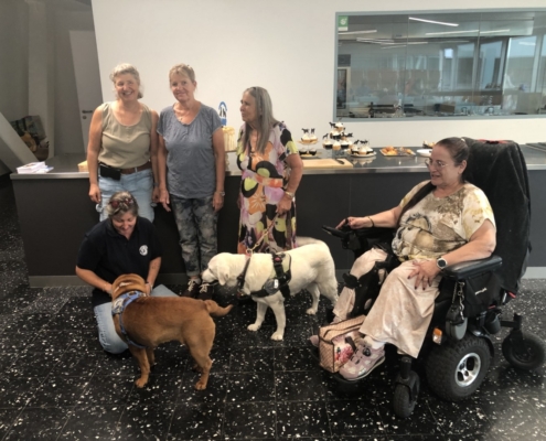 Photo de groupe avec cinq dames et deux chiens d’assistance devant un comptoir garni de gâteaux et cupcakes.