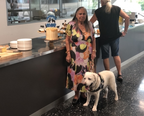 Une dame debout avec un chien guide devant un gâteau d’anniversaire décoré « 20 ans », sur un buffet dans une salle lumineuse.