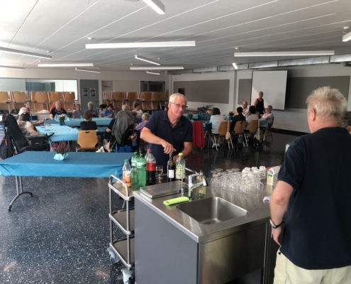 Un personne sert des boissons à des convives assis autour de tables bleues lors d’un moment convivial dans une grande salle.