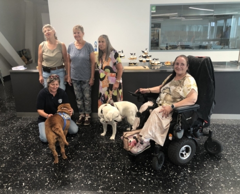 Photo de groupe avec cinq dames et deux chiens d’assistance devant un comptoir garni de gâteaux et cupcakes.
