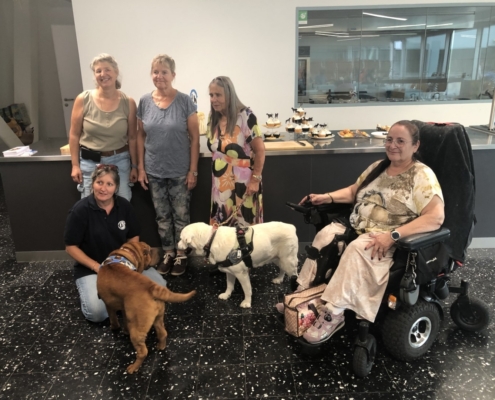 Photo de groupe avec cinq dames et deux chiens d’assistance devant un comptoir garni de gâteaux et cupcakes.