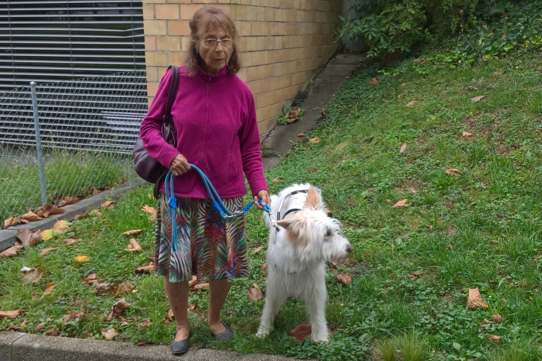 Une dame âgée tenant son chien d’assistance Nino, debout sur une pelouse près d’un bâtiment en briques.