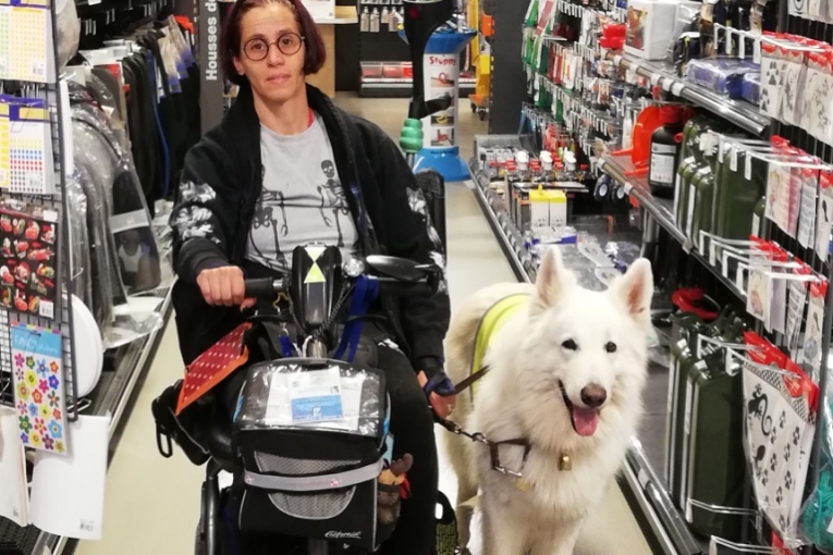 Dame en fauteuil roulant accompagnée de son chien d’assistance blanc dans une allée de magasin.