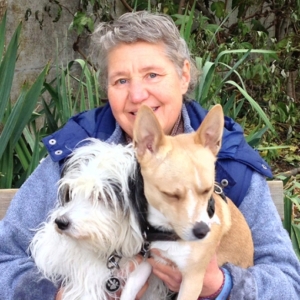 Dame souriante assise sur un banc, tenant deux petits chiens dans ses bras, entourée de verdure en arrière-plan.