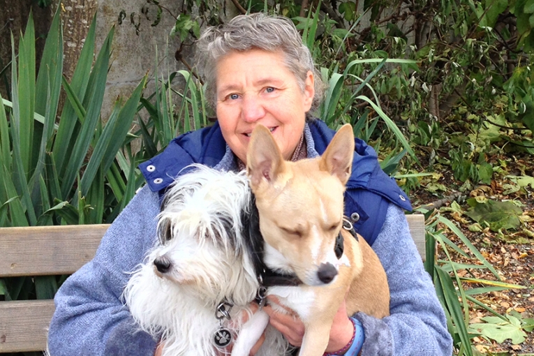 Dame souriante assise sur un banc, tenant deux petits chiens dans ses bras, entourée de verdure en arrière-plan.