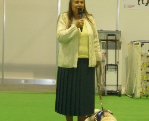 Une dame tenant un micro aux côtés de son chien d’assistance allongé, lors d’une présentation.