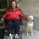 Dame souriante en fauteuil devant une entrée, accompagnée de son chien guide, assis à ses côtés.