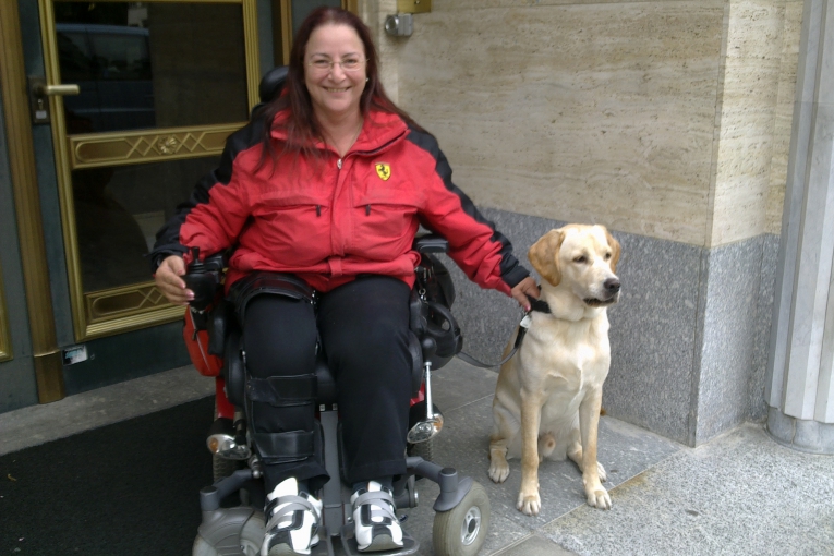 Dame souriante en fauteuil devant une entrée, accompagnée de son chien guide, assis à ses côtés.