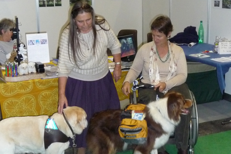 Deux dames accompagnées de leurs chiens d’assistance se rencontrent lors d’un événement, souriantes et entourées de stands.