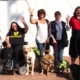 Six dames posent avec leurs chiens d’assistance, souriantes et la main levée, sous un beau soleil.