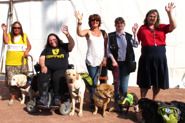 Six dames posent avec leurs chiens d’assistance, souriantes et la main levée, sous un beau soleil.