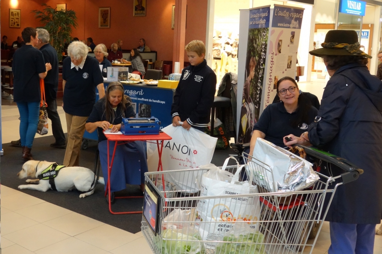 Stand L’Autre Regard dans un centre commercial, avec un chien-guide, plusieurs dames de l'association et des échanges avec les passants.
