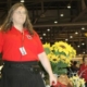 Une dame en chemise rouge et jupe noire, badge autour du cou, se tient debout dans un salon animé.