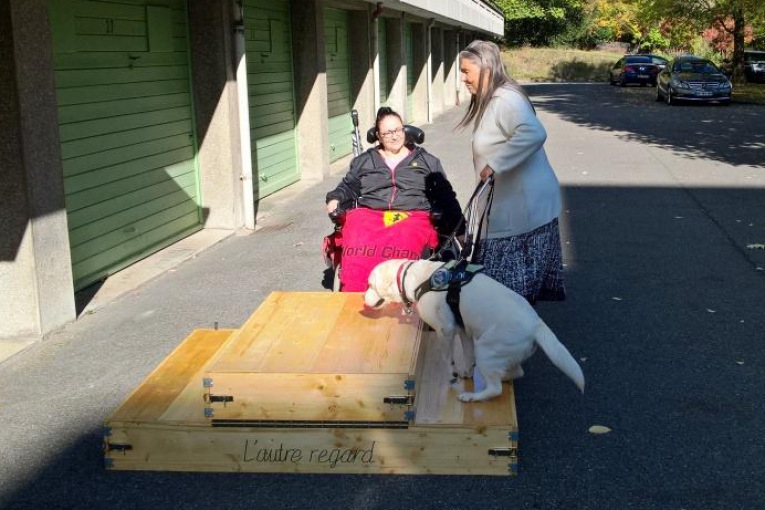 Démonstration d’un chien guide montant une rampe en bois, observé par deux femmes dont l’une en fauteuil roulant.