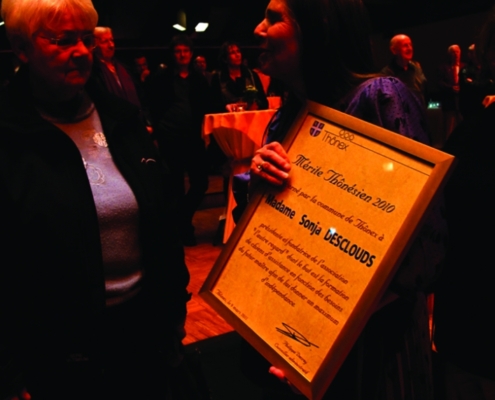 Une dame reçoit un diplôme honorifique encadré, intitulé "Mérite Thônésien 2010", lors d'une cérémonie.