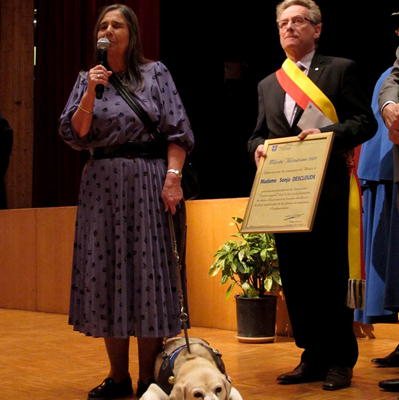 Une dame prend la parole sur scène, accompagnée de son chien-guide allongé, tandis qu’un élu lui remet un diplôme honorifique.