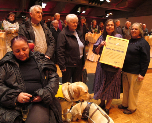 Une dame pose avec un diplôme honorifique entourée de proches et de deux chiens-guides.