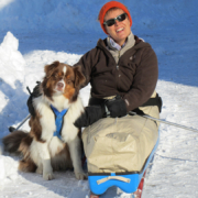 Une dame souriante en luge adaptée, assise dans la neige aux côtés de son chien d’assistance Shantih, un berger australien.