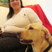 Une dame en fauteuil roulant sourit, assise à côté de son chien d’assistance Sorbonne, attentif et calme.