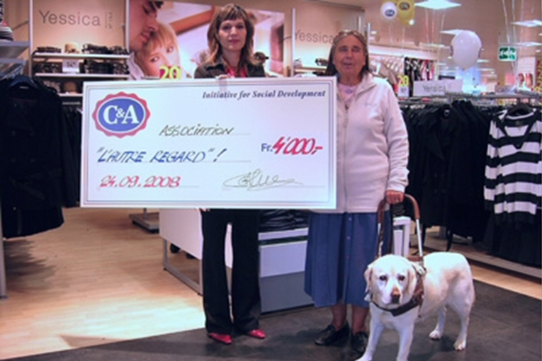 Remise d’un chèque de Fr. 4’000.– à l’association L’Autre Regard, au nom de C&A, en présence d’une dame avec son chien guide.